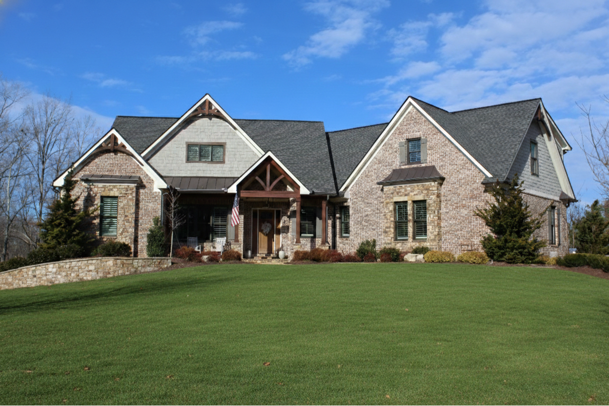 A large suburban home with stone and light-colored siding, a dark roof, and a well-manicured lawn under a sunny blue sky.