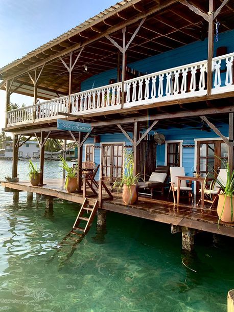 Waterfront wooden house with balcony, ladder, and chairs over clear green water