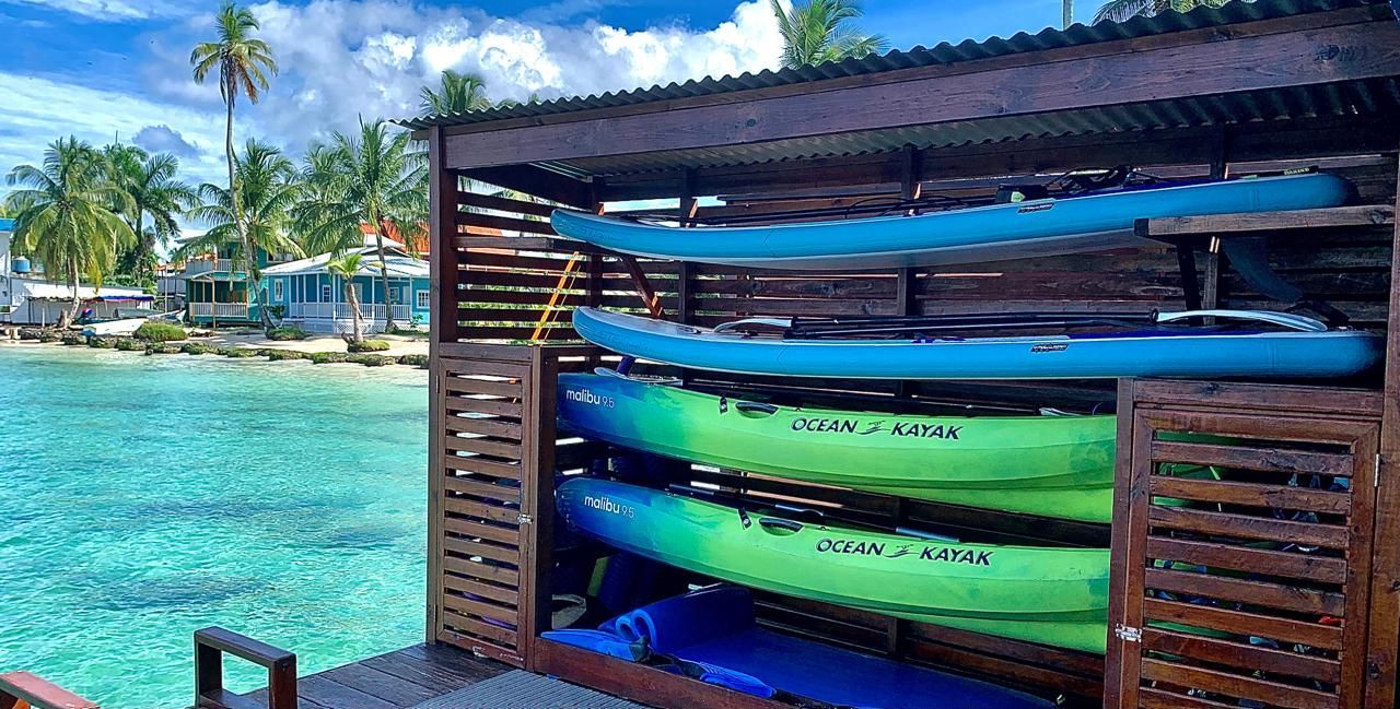 Colorful kayaks stacked in a wooden rack by a turquoise tropical beach with palm trees and blue sky.