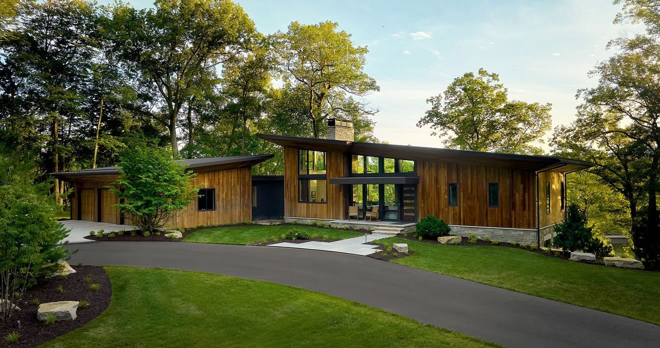 Modern wooden house with large windows, surrounded by trees and a curved driveway in a green yard