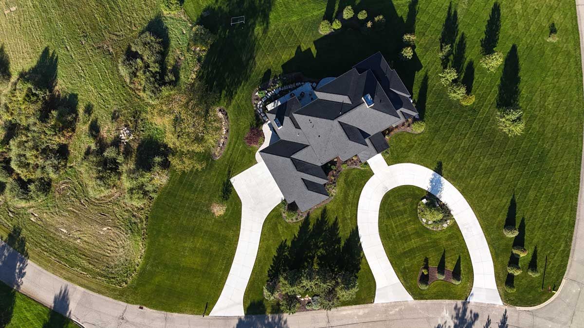 Aerial view of a house with a curved driveway, green lawn, and scattered trees.