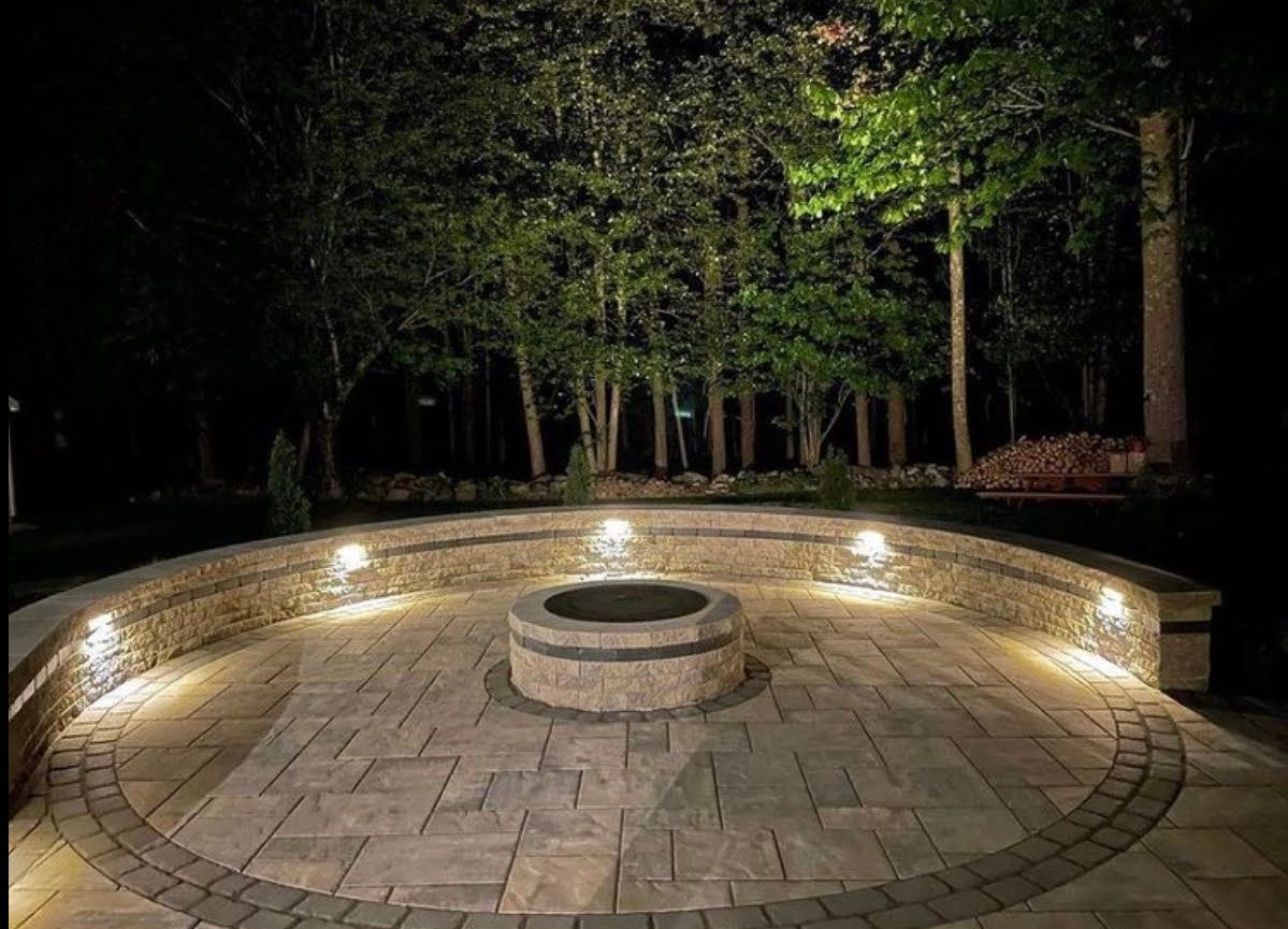 Circular stone patio with a central fire pit, lit at night among trees
