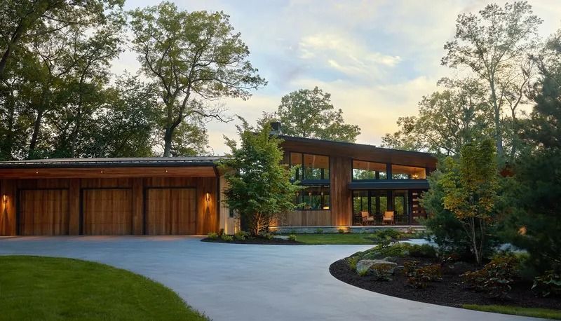 Modern house with wooden exterior, driveway, and lush landscaping against a backdrop of trees.