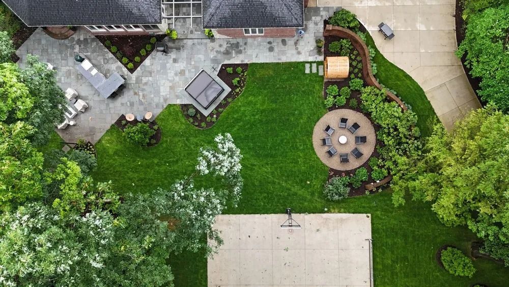 Aerial view of a landscaped backyard with a house, patio, fire pit, basketball court, and lush greenery.