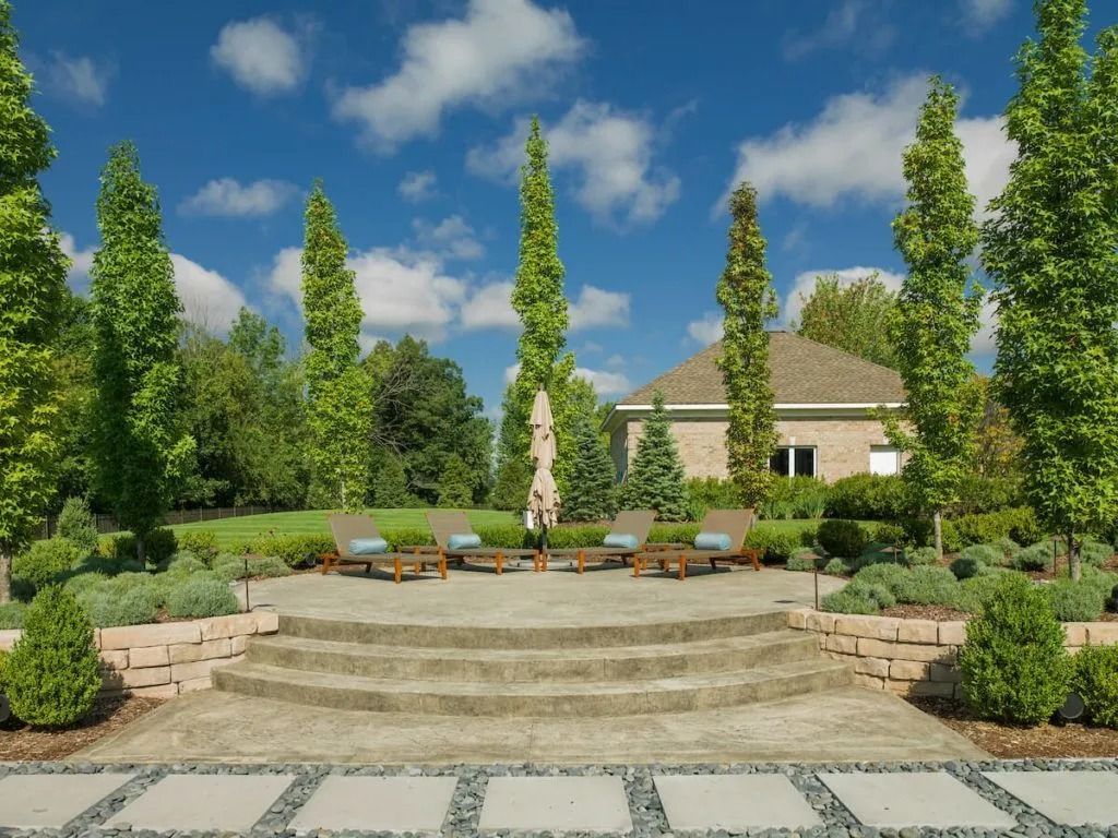 Stone patio with lounge chairs, surrounded by landscaping and tall trees, a house in the background.