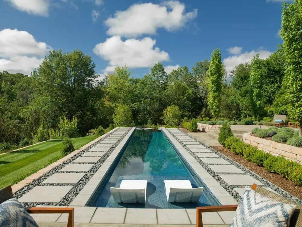 Long, rectangular pool with white lounge chairs, surrounded by landscaping and trees.