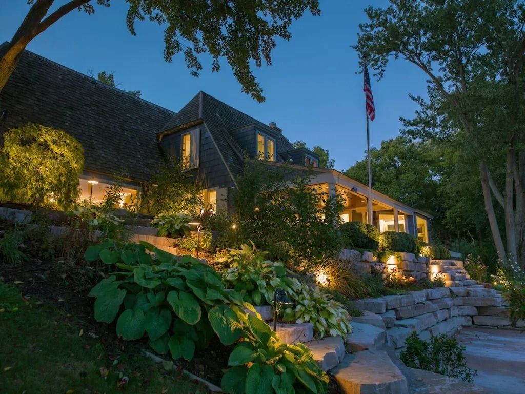 House at dusk with lit landscaping, flag, and green foliage.