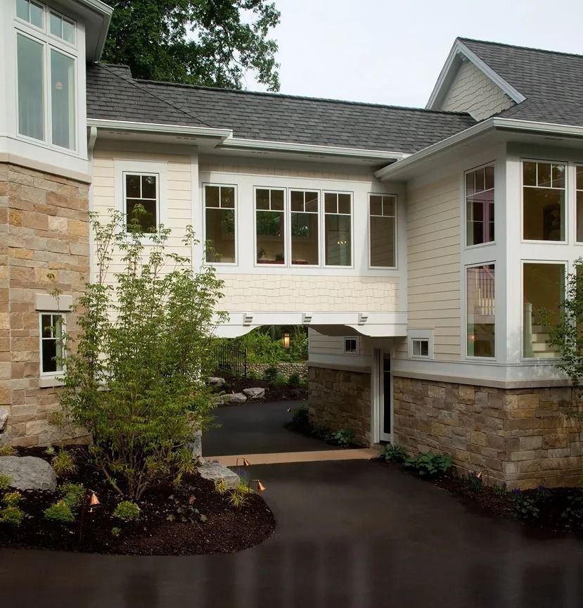Beige and stone home with a bridge-like structure over a dark driveway, trees in the background.