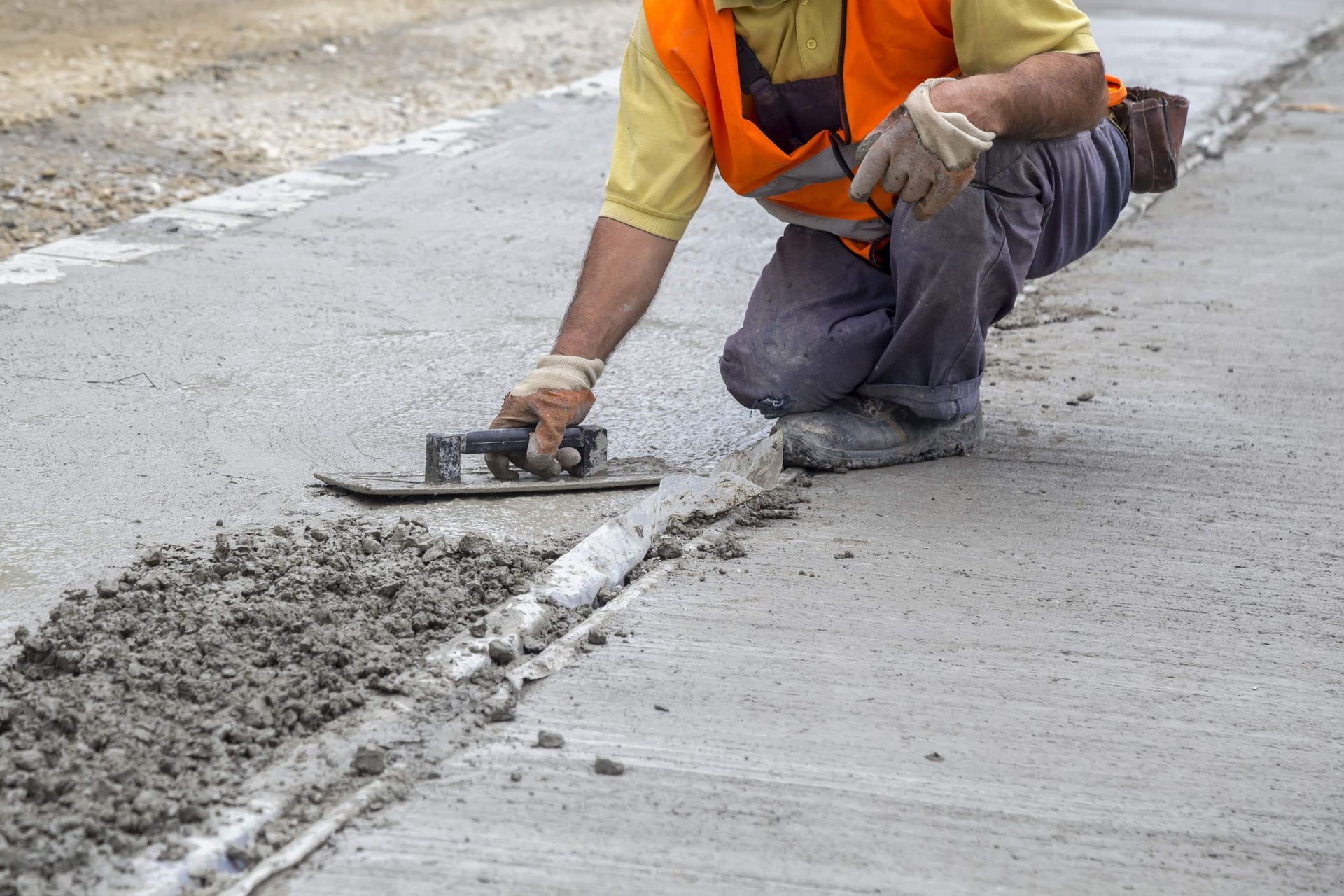 Worker in orange vest kneels, smoothing concrete with a trowel along a road joint.