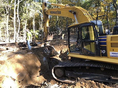Yellow excavator digging in a dirt area, with a worker standing nearby in a wooded setting.
