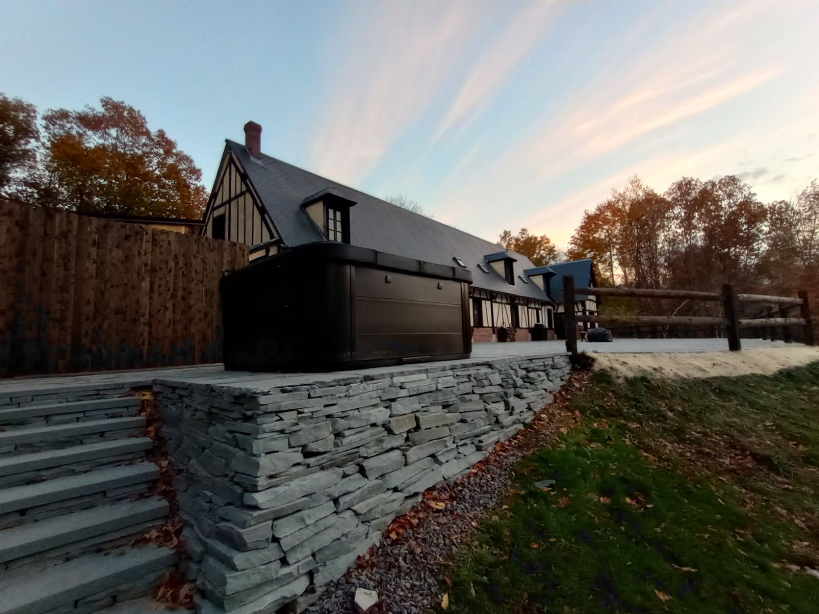 Stone staircase leads to a dark, modern hot tub and a building with a slate roof in a natural setting.
