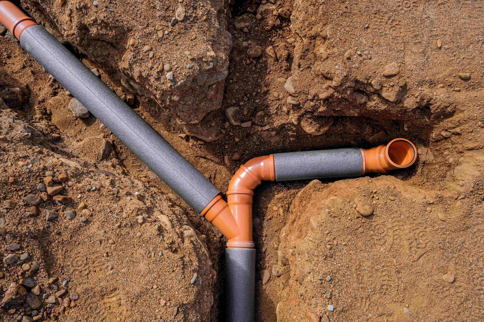 Orange and gray pipes and fittings in a trench, buried in sand and soil.
