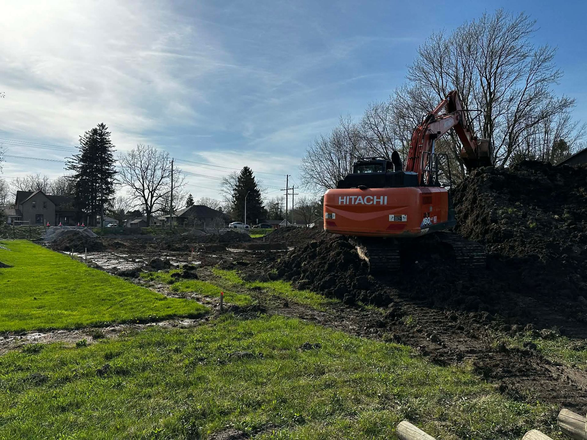 Hitachi excavator clearing muddy ground beside a green field under a bright sky