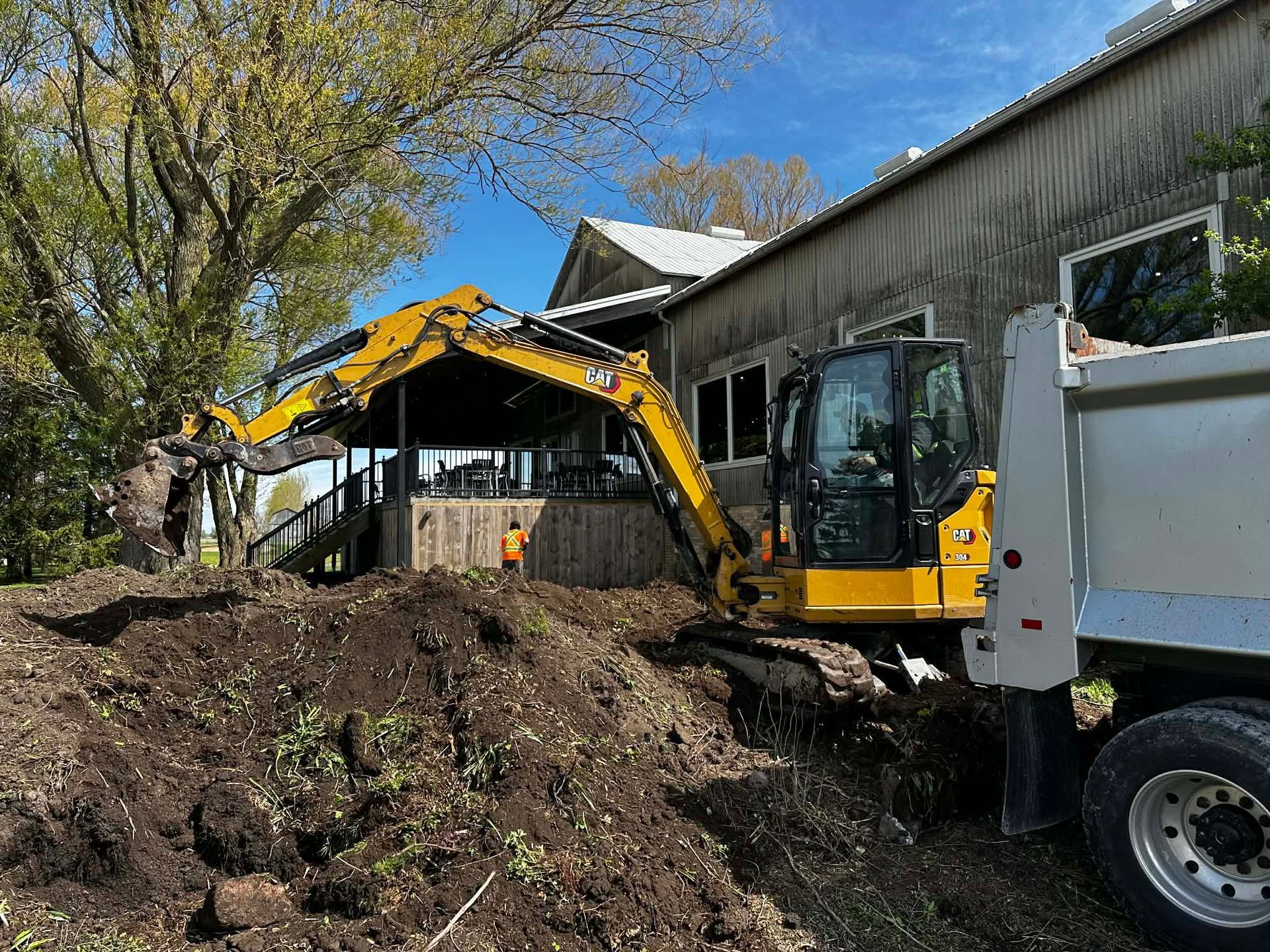 Yellow excavator loading dirt into a white dump truck beside a building and trees