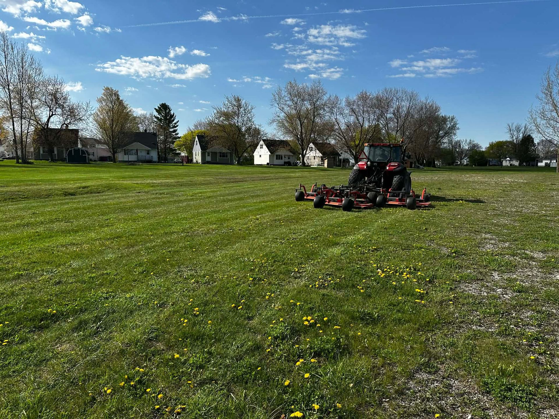 Person mowing a grassy field under a blue sky, with houses and trees in the background.