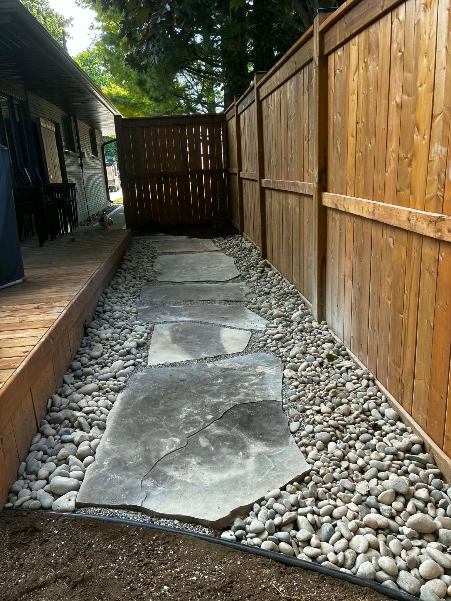 Stepping stone path beside a wooden fence, bordered by gray river rocks along a narrow side yard.
