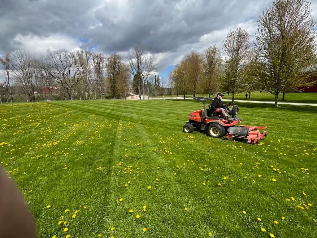 Person mowing a green field with a red riding mower under cloudy skies