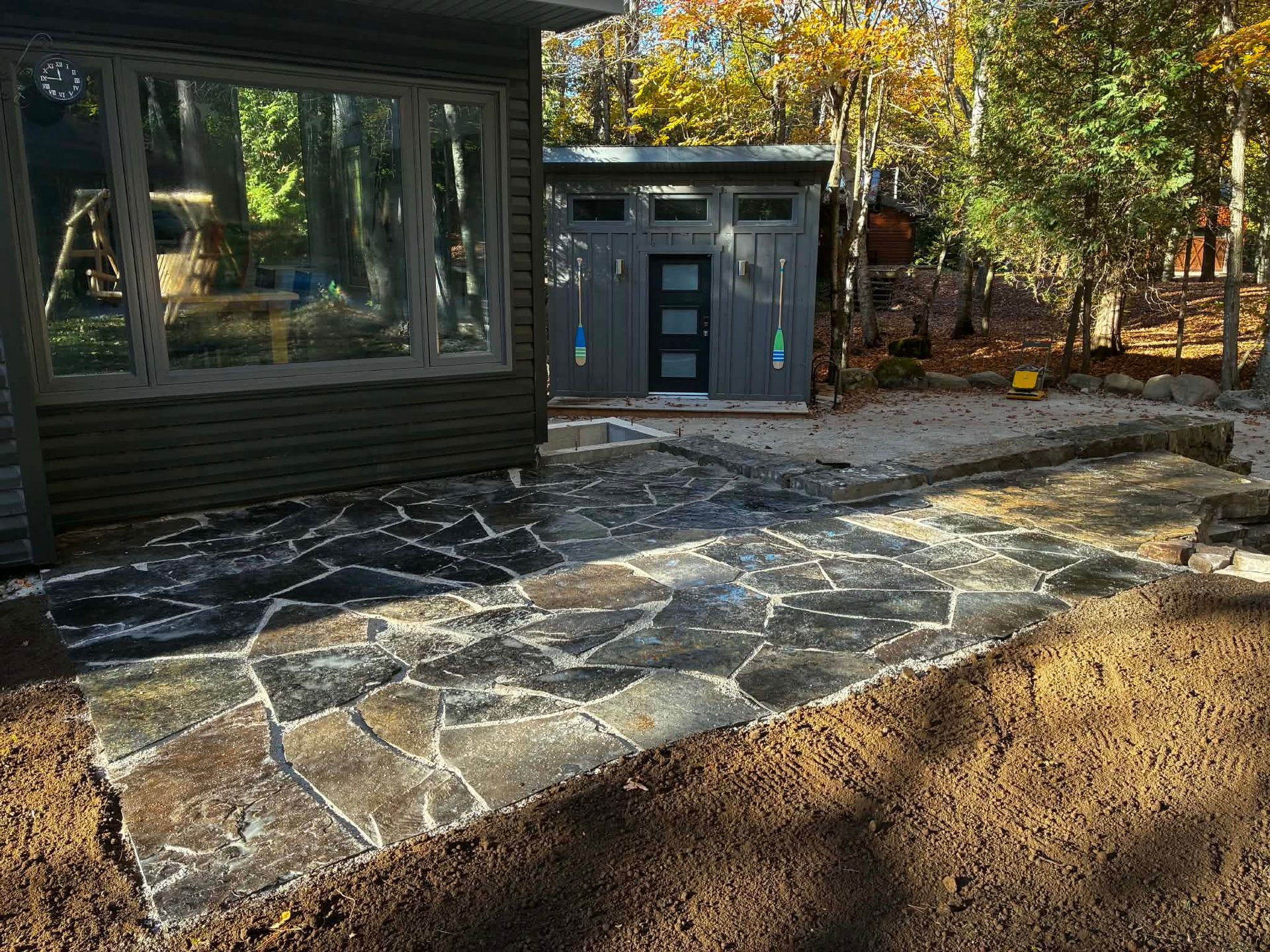 Stone patio beside a dark house and shed, with a dirt foreground and autumn trees in back.