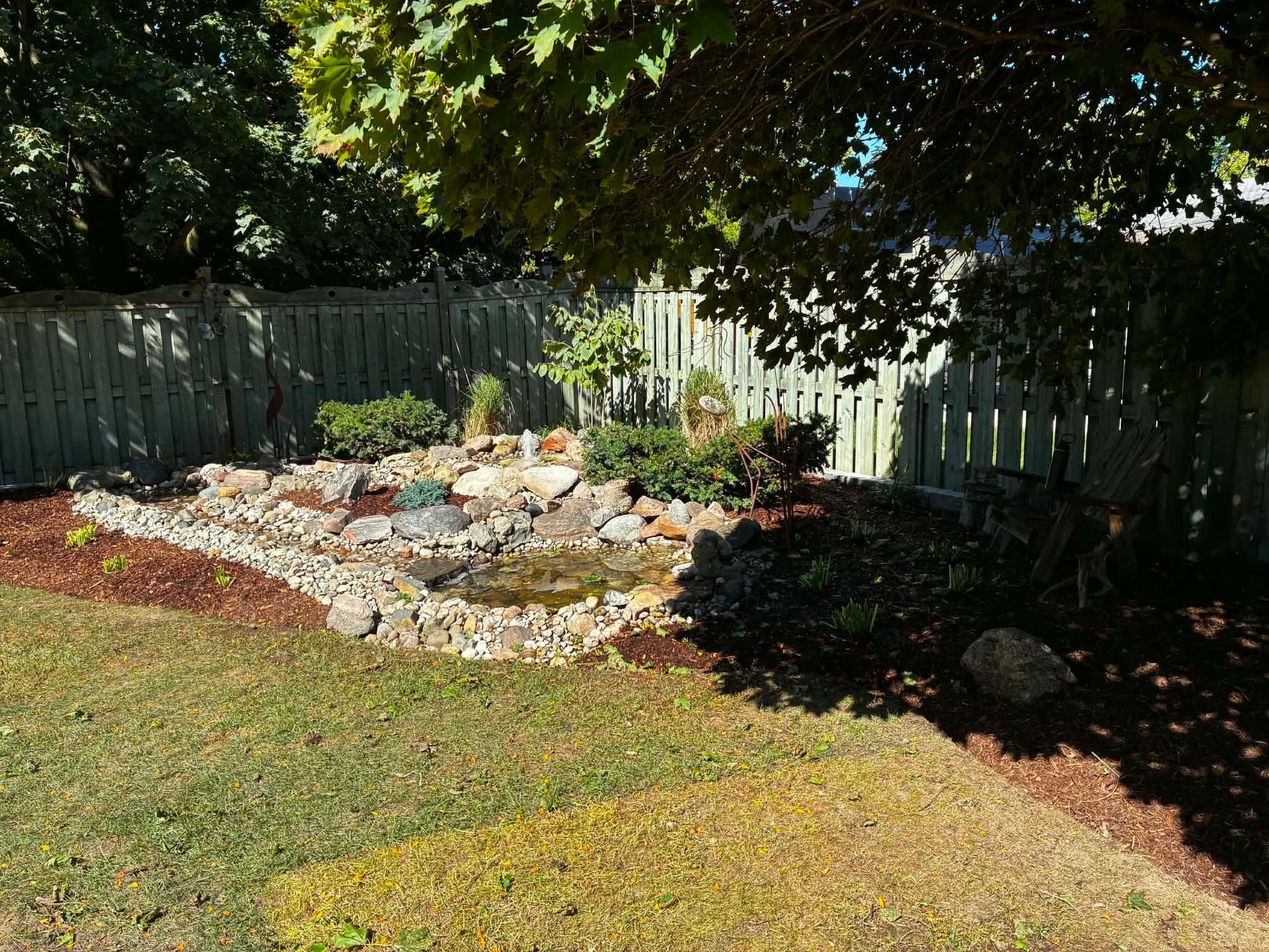Backyard rock garden with flowers, bordered by a white fence under a leafy tree