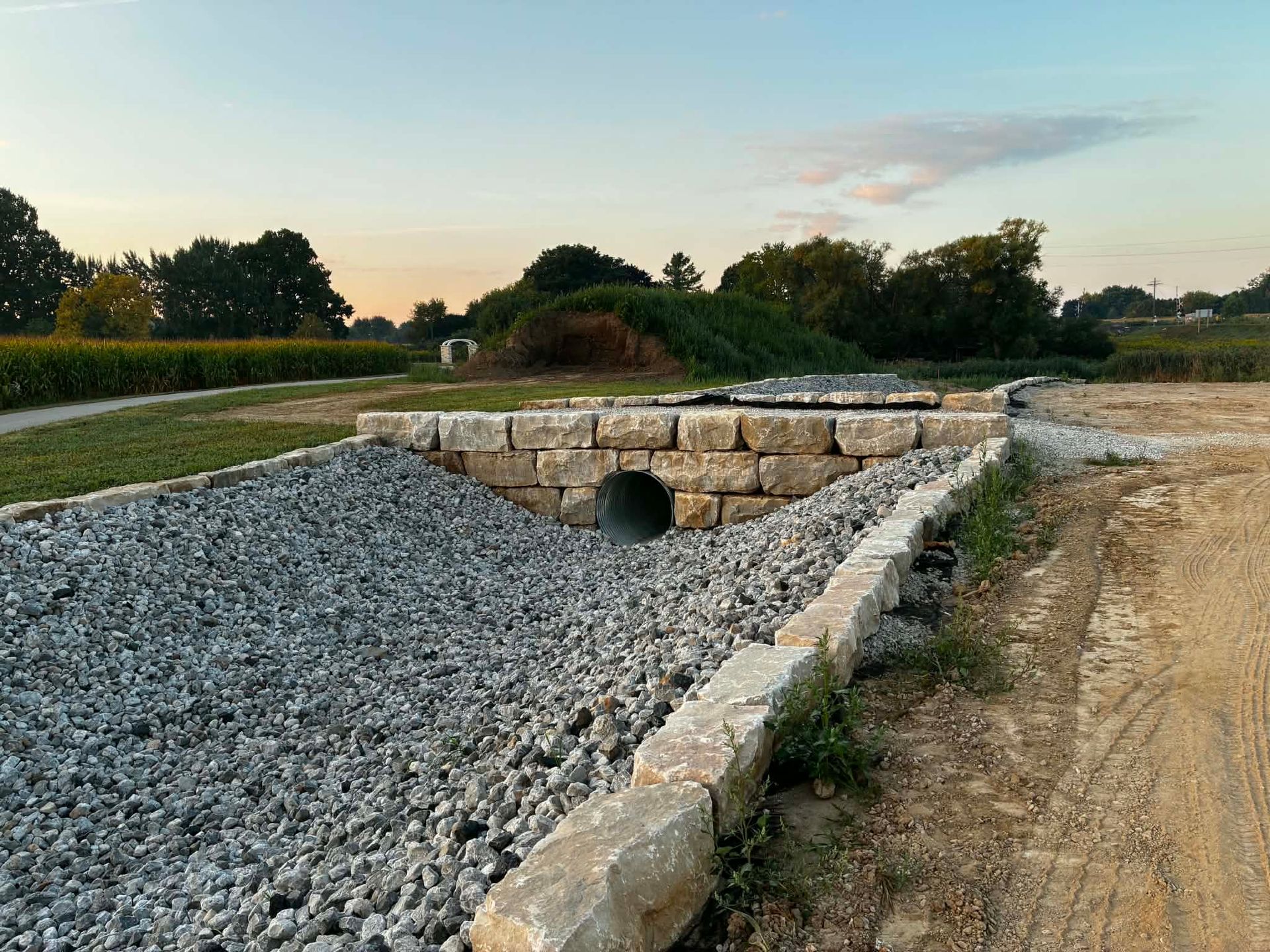 Stone-lined drainage ditch beside a gravel path at sunset, with grassy fields and trees in the background