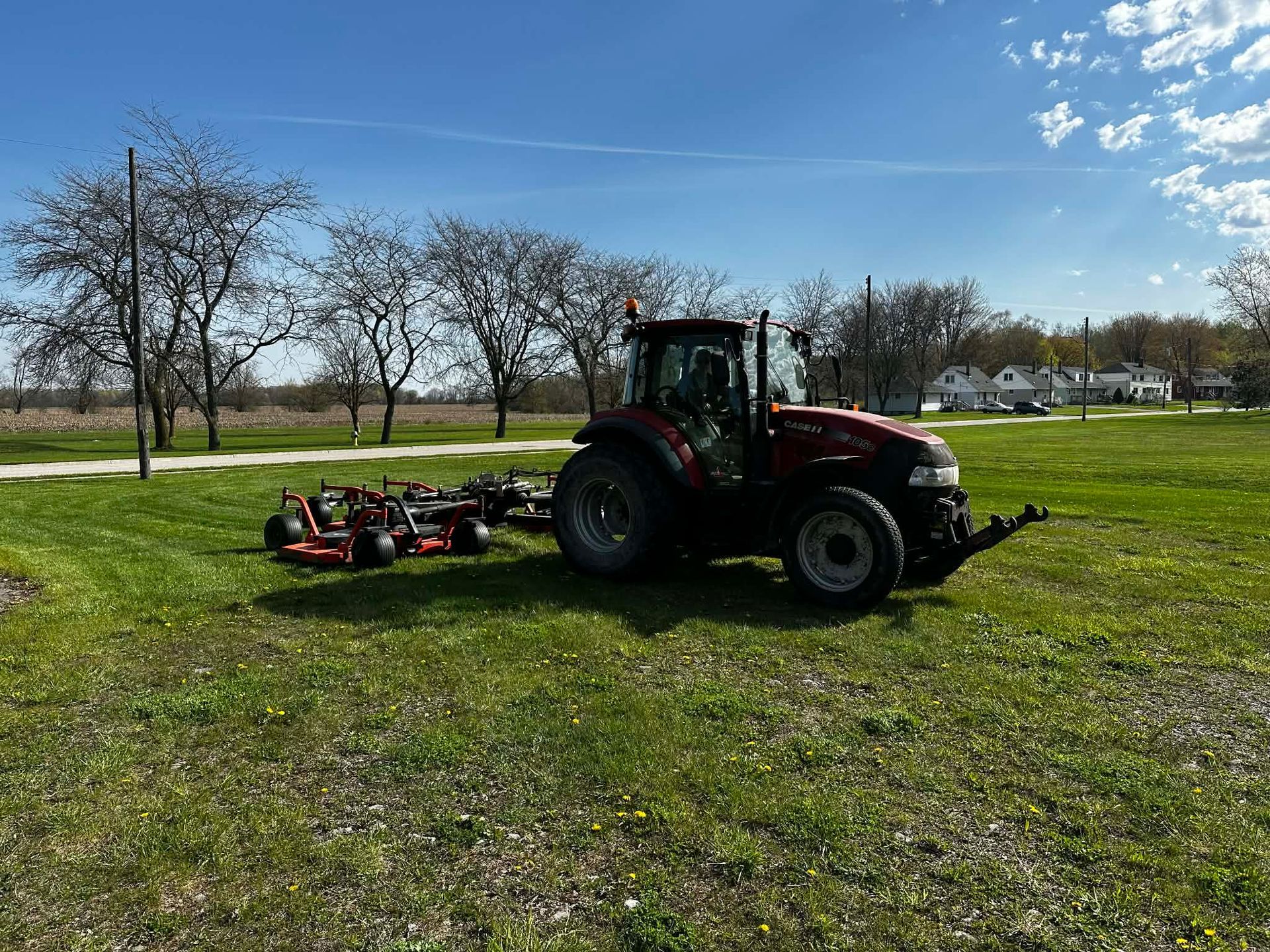 Person mowing a grassy field under a blue sky, with trees and houses in the background