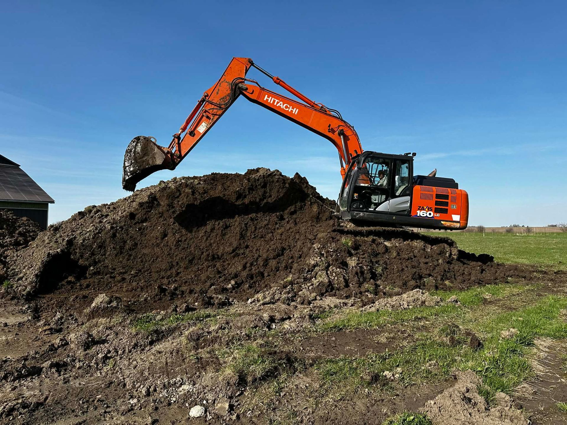 Hitachi excavator dumping dirt into a large pile in a grassy field under a clear blue sky