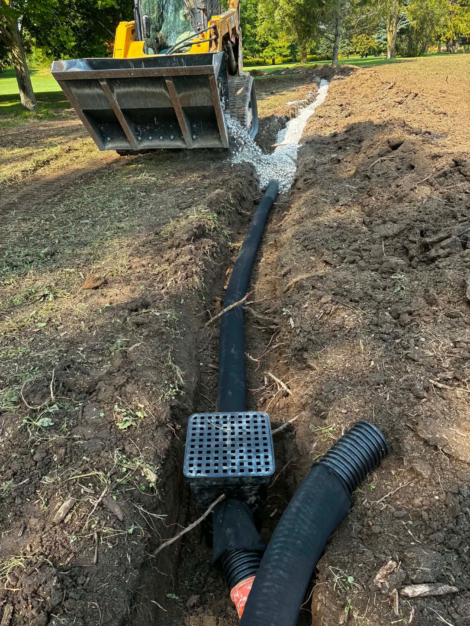 Excavator bucket beside a black drainage pipe laid in a dirt trench on a construction site