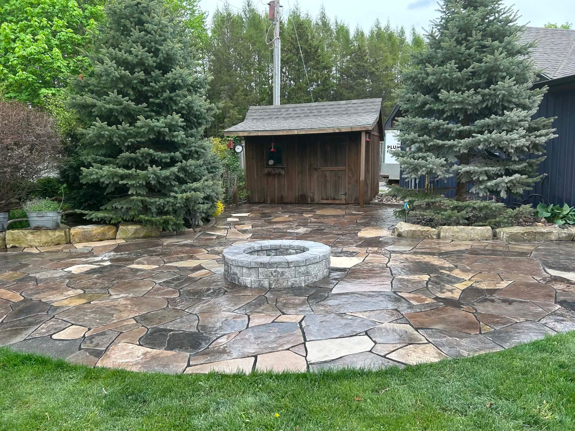 Stone patio with circular fire pit and small wooden shed, surrounded by evergreen trees and landscaping.