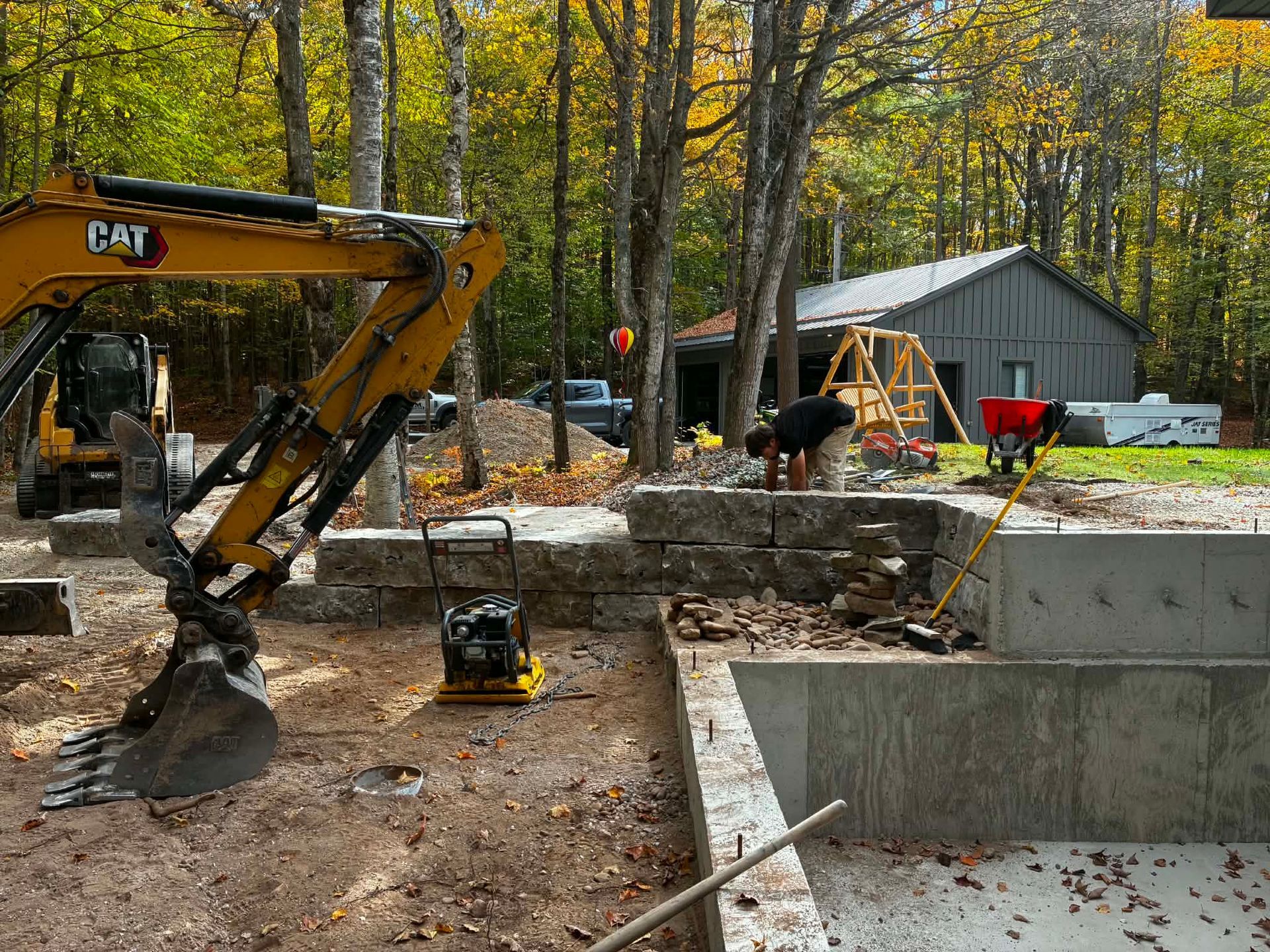 Construction site with excavator beside a concrete foundation in a wooded yard