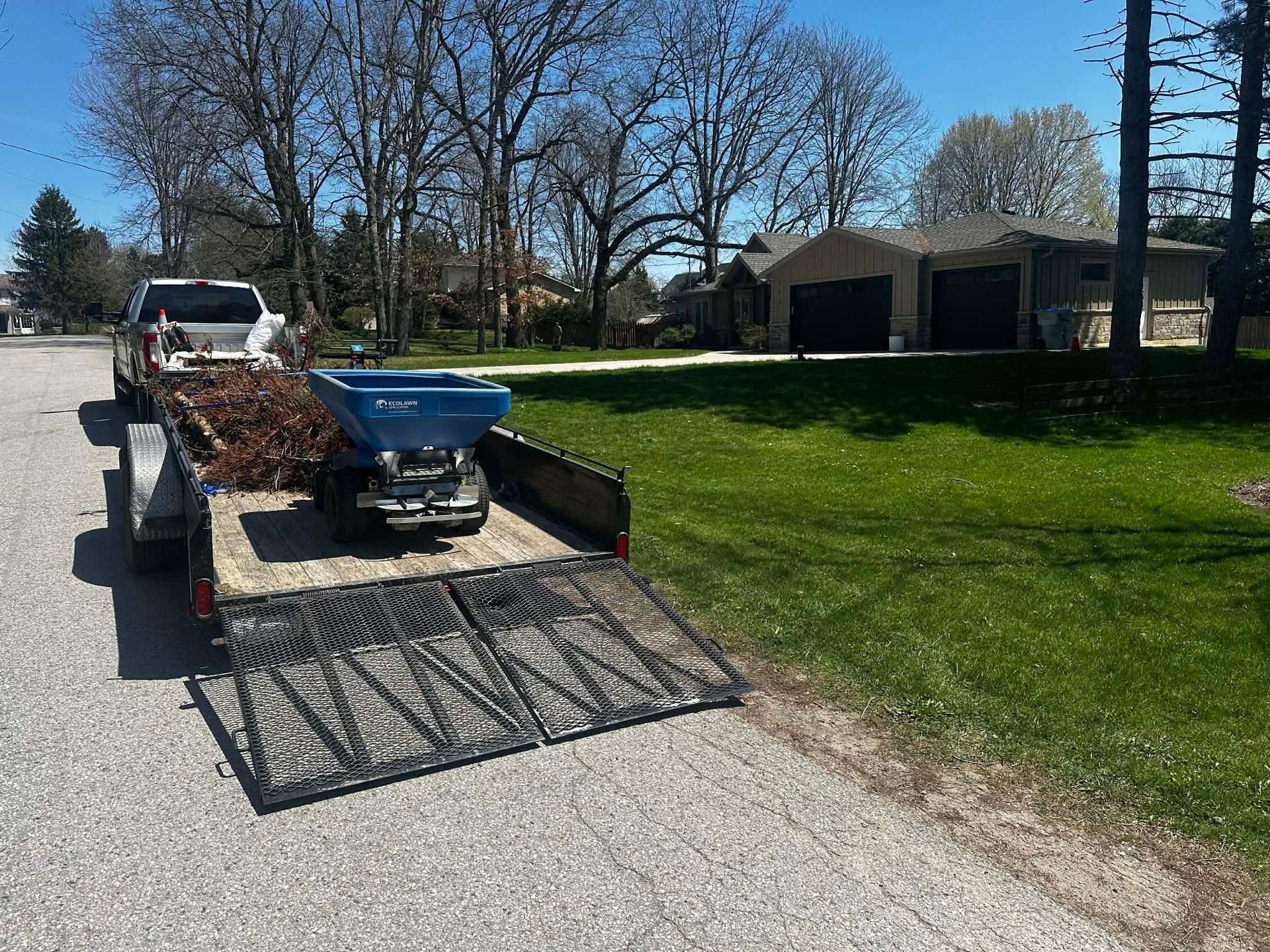 Utility trailer loaded with branches and a wheelbarrow, parked on a suburban street beside a lawn