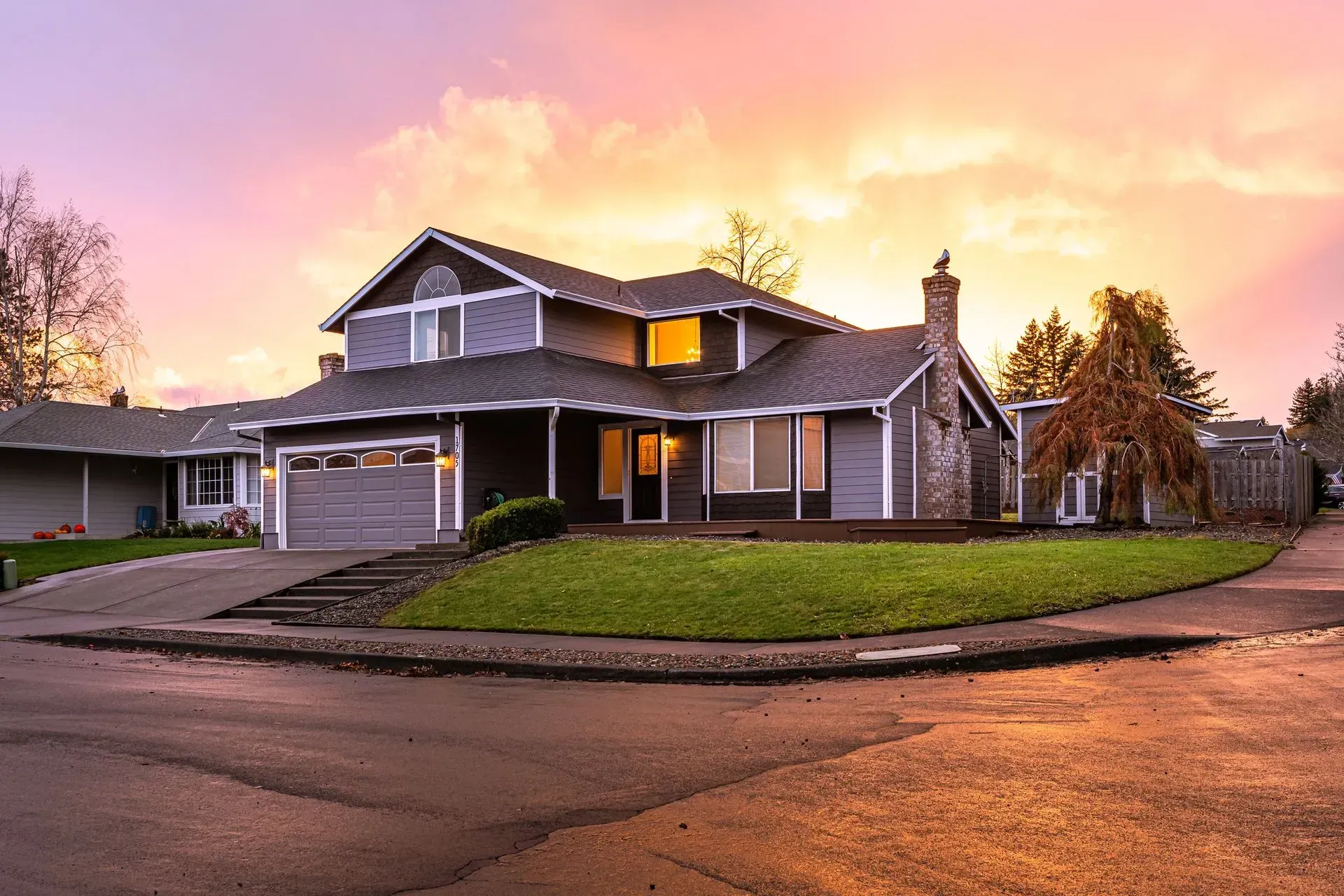 Suburban house at sunset with warm lights, a garage, and a curved driveway under a pink-orange sky