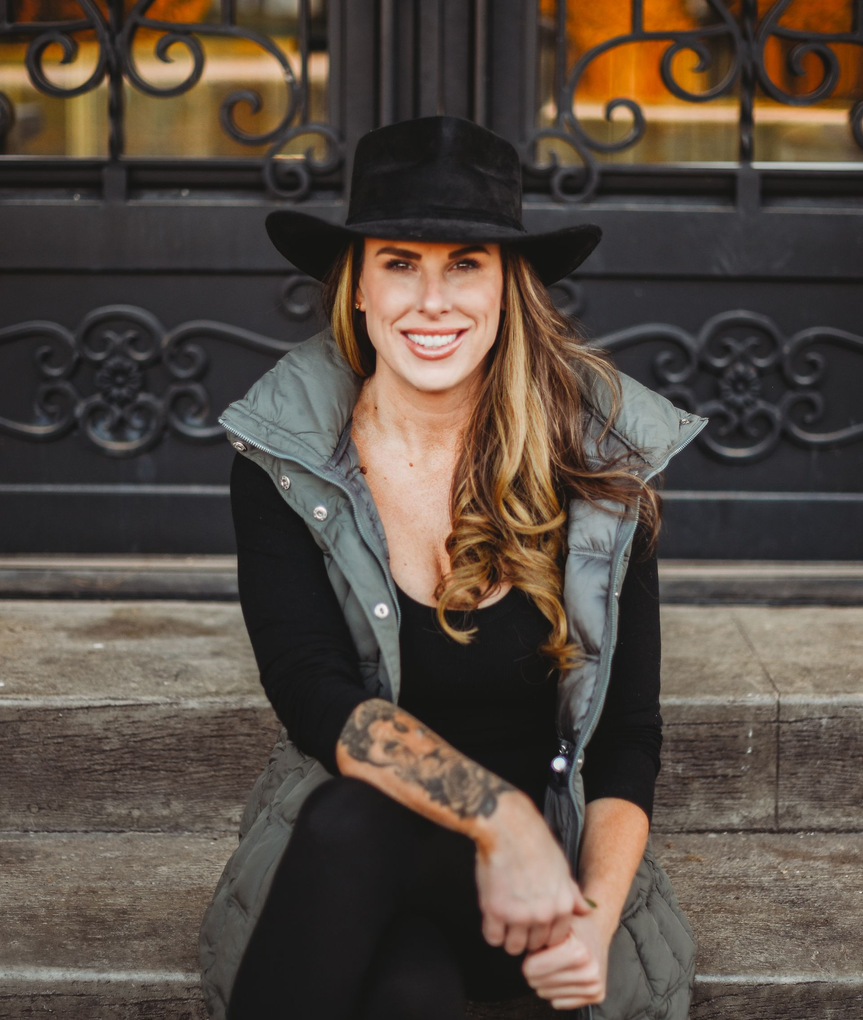 Smiling woman in a black hat and vest, seated against a dark background.