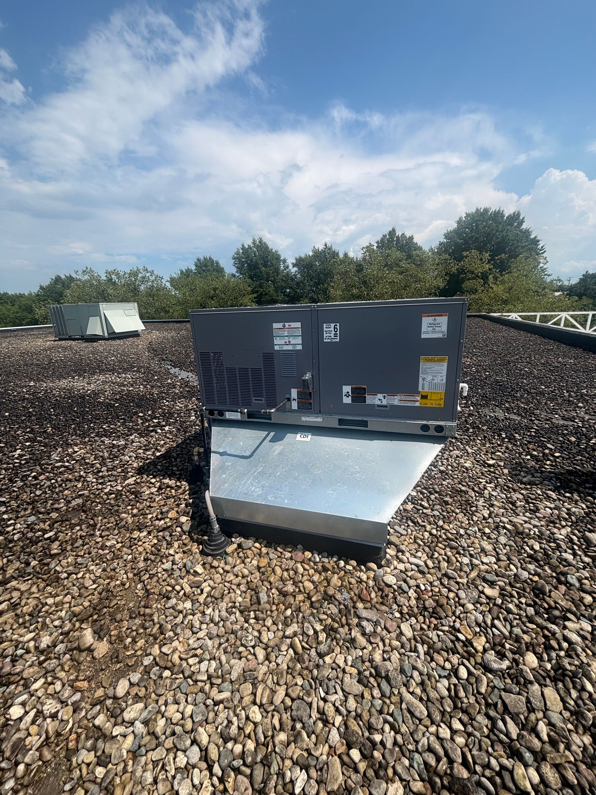 HVAC unit on a pebble-covered rooftop under a bright blue sky with trees in the background.