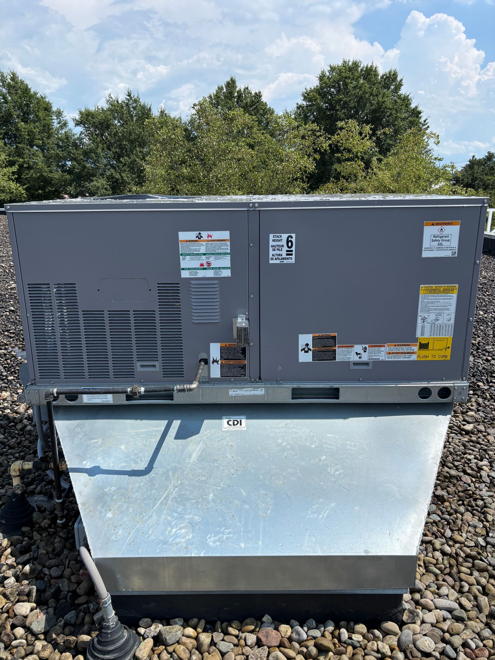 Gray rooftop HVAC unit on a silver metal base, on a gravel roof, with trees in the background under a blue sky.