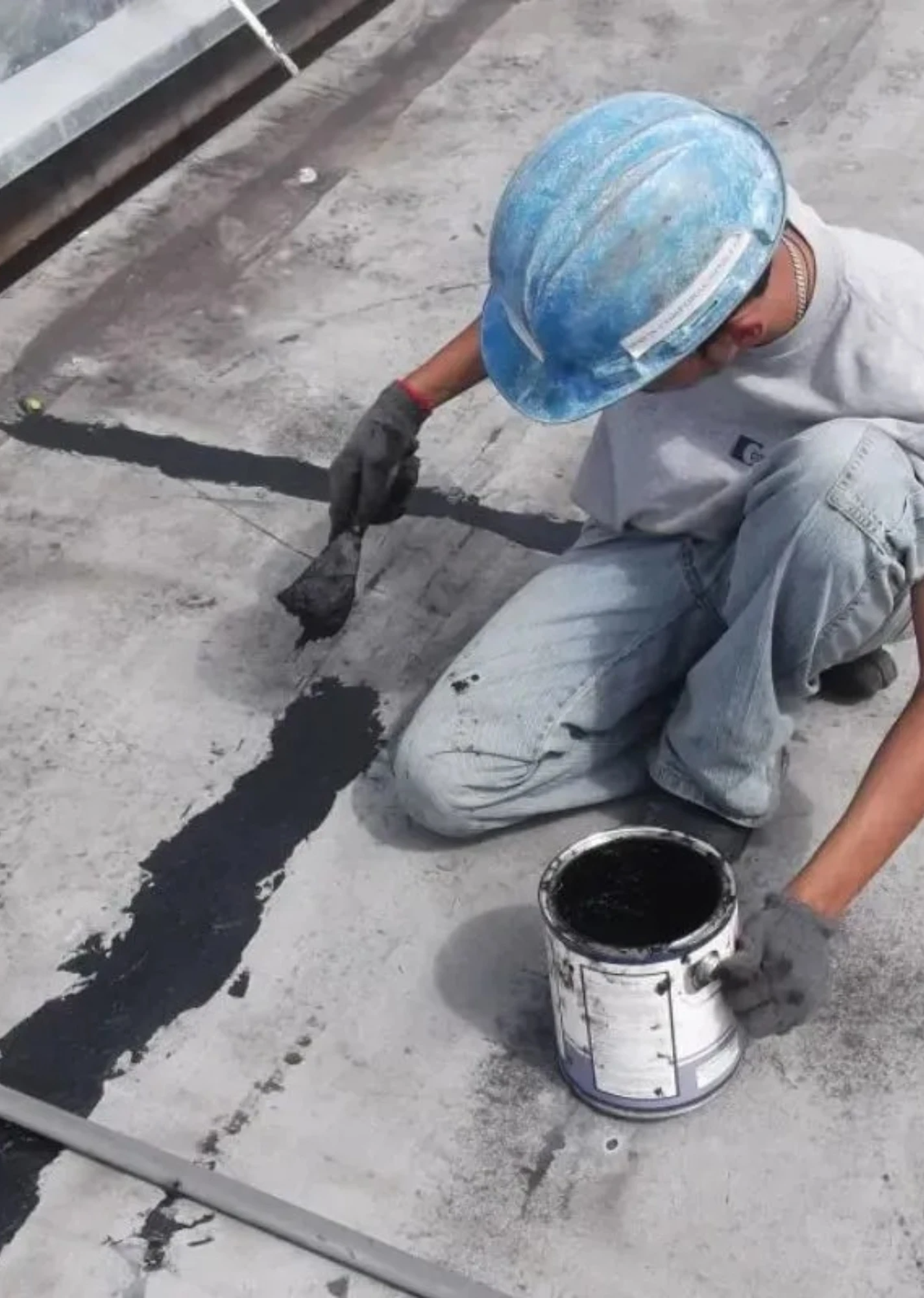 Un trabajador con casco azul aplica una capa impermeabilizante negra a una superficie de hormigón con una brocha.