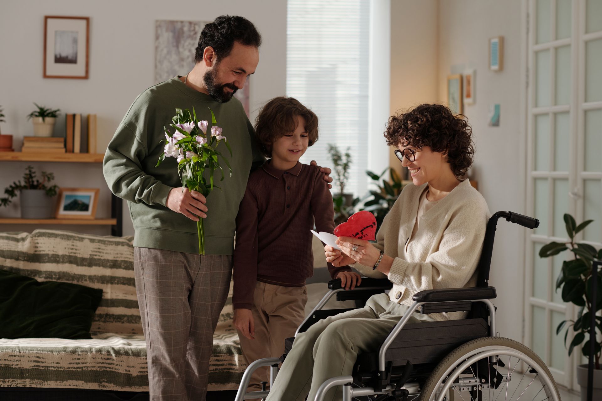 Three people in a living room, one in a wheelchair, smiling at a bouquet of flowers and a card.