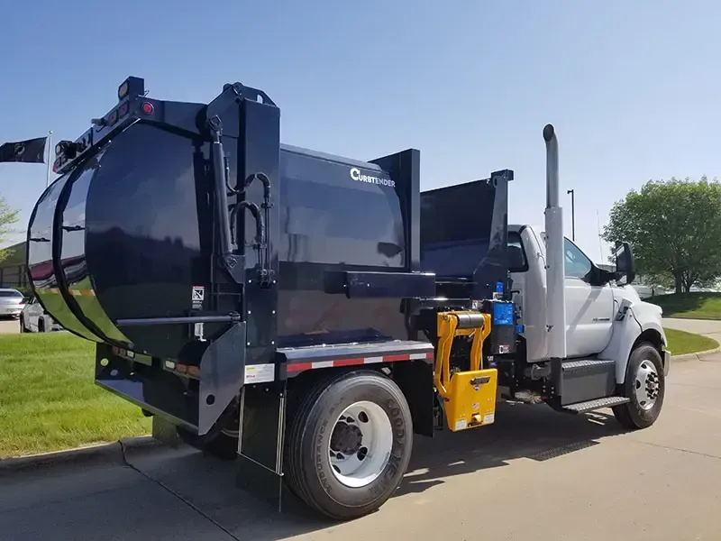 Black garbage truck on a road beside a grassy field, with the cab door open.