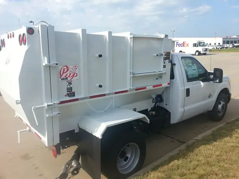 White waste collection truck parked on grass beside a building, side view with red logo and black rear bin.