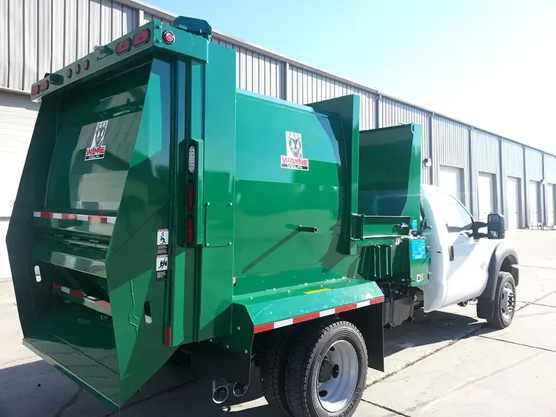 Green garbage truck with open rear hopper parked beside a white truck trailer.