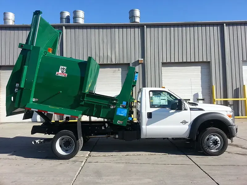 White dump truck with green raised bed parked outside a warehouse under bright sunlight
