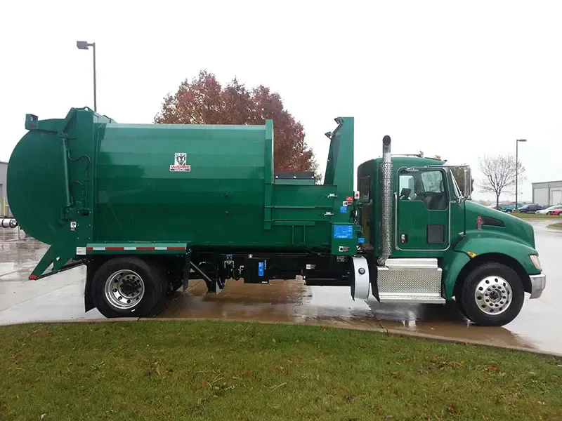 Green garbage truck parked on wet pavement beside a grassy area