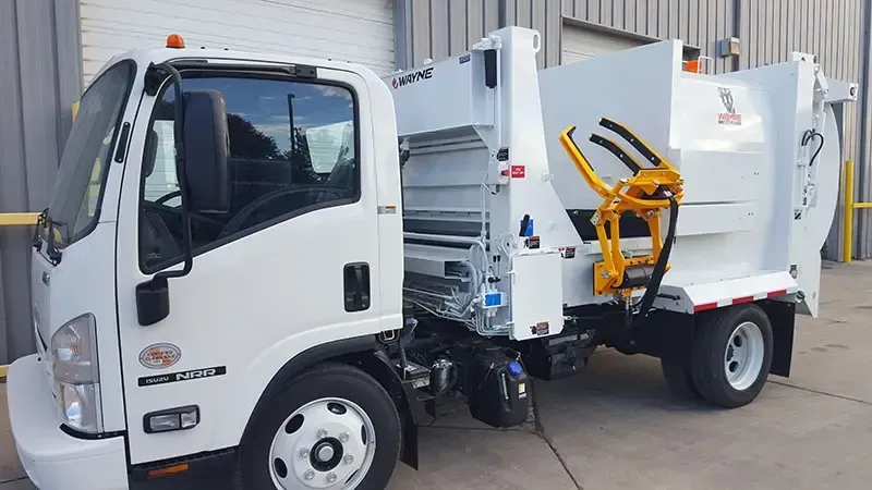 White compact garbage truck with yellow lift mechanism parked outdoors beside a warehouse