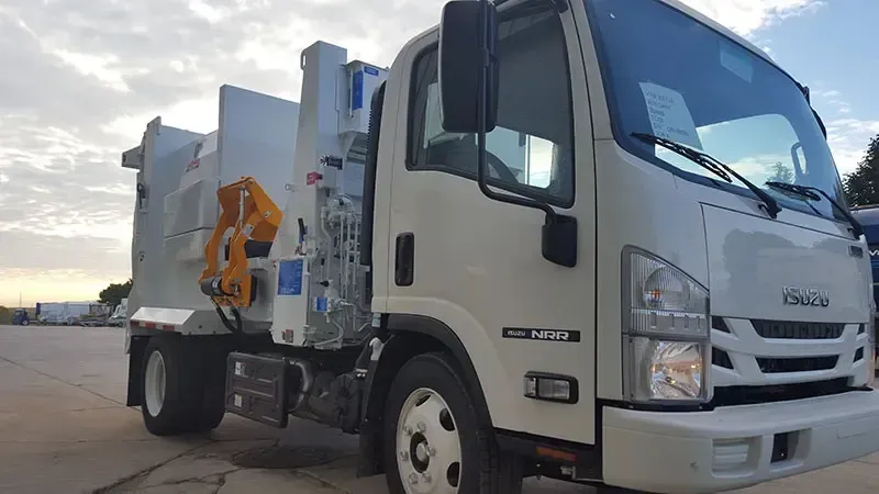 White Isuzu utility truck with an orange lifting arm parked on a concrete lot at sunset