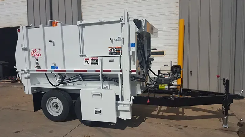White utility trailer with mounted equipment and black boom on a concrete lot outside a warehouse