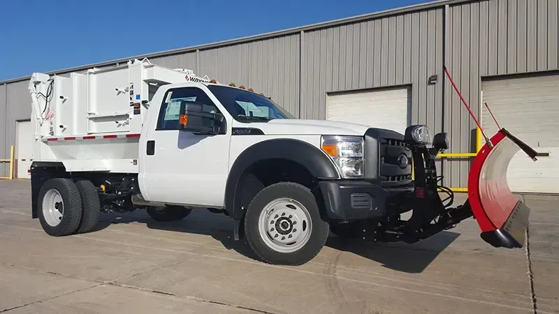 White utility truck with a red snowplow attachment parked outside a beige warehouse.