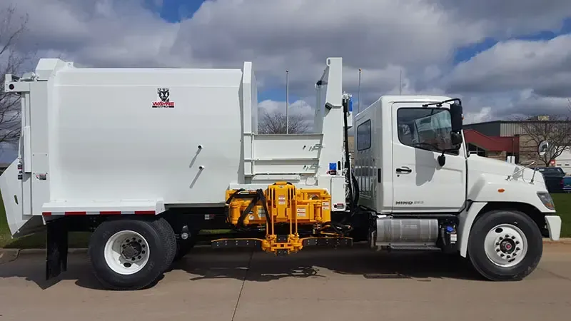 White garbage truck with side-loading arm parked on a street under a cloudy sky