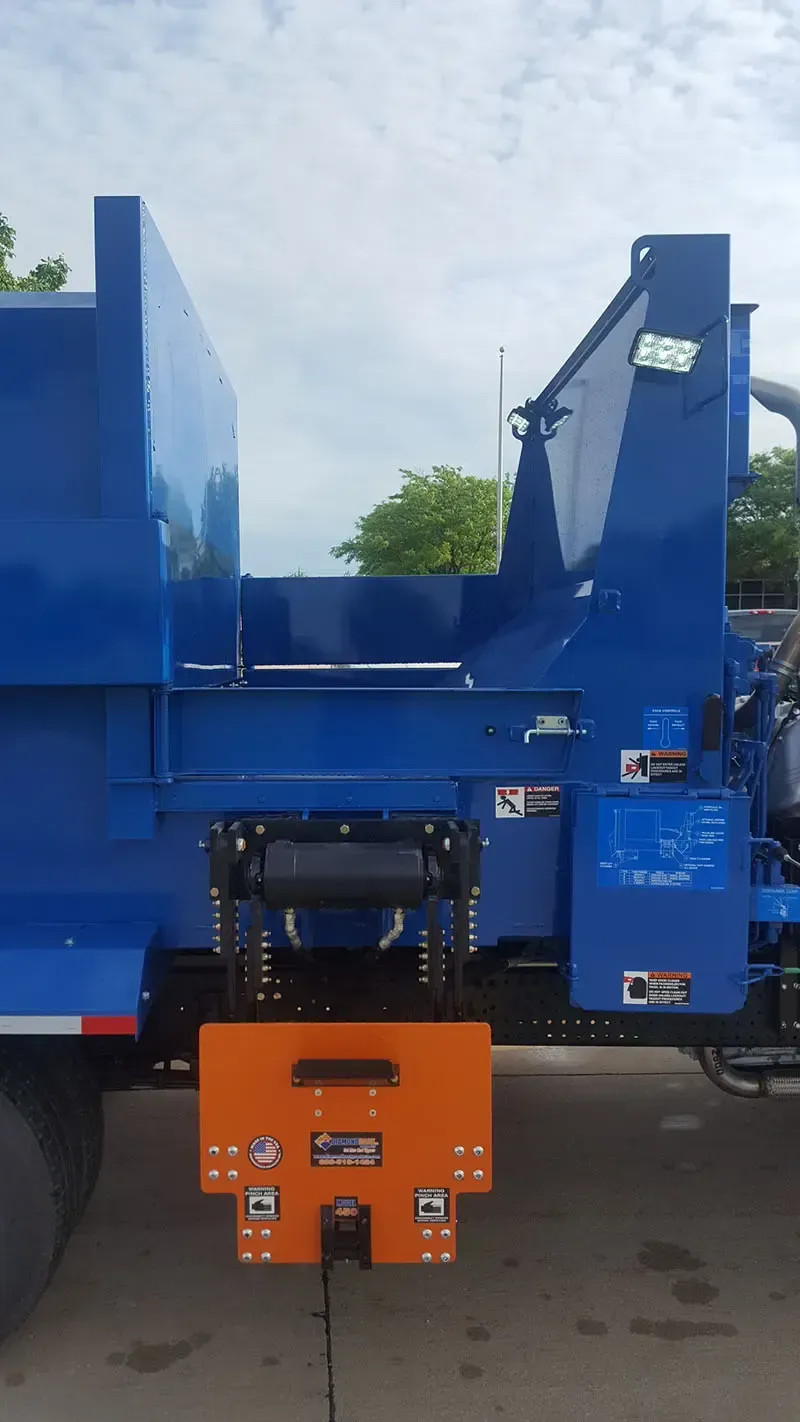 Blue industrial truck with a blue hopper and orange rear hitch attachment, parked outdoors.