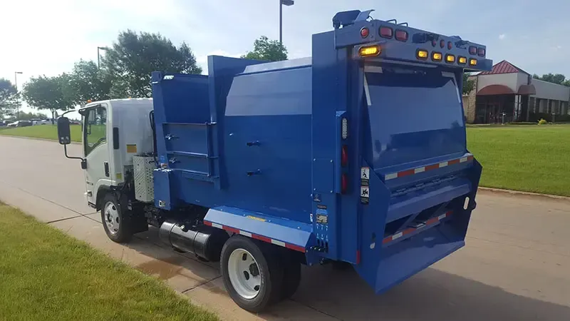 Blue and white garbage truck on a residential street, viewed from the rear side.