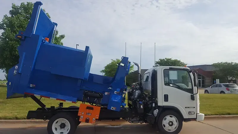 Blue garbage truck lifting a large dumpster on a street beside a grassy area