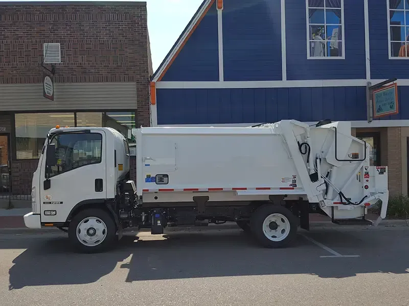 White garbage truck parked on a city street beside brick and blue buildings.